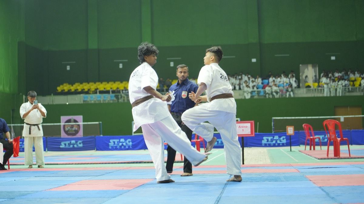 Two karateka in white gi exchange kicks at a KKFI tournament while the referee watches